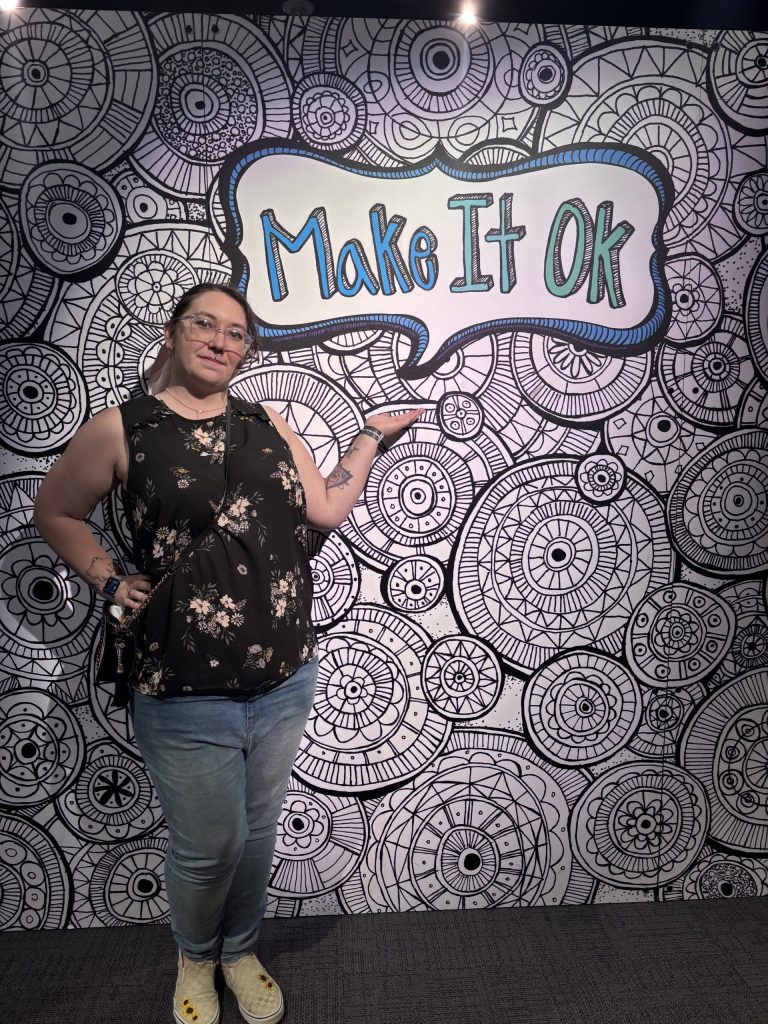 Lexi Lucas stands in front of rural that says "Make it OK"